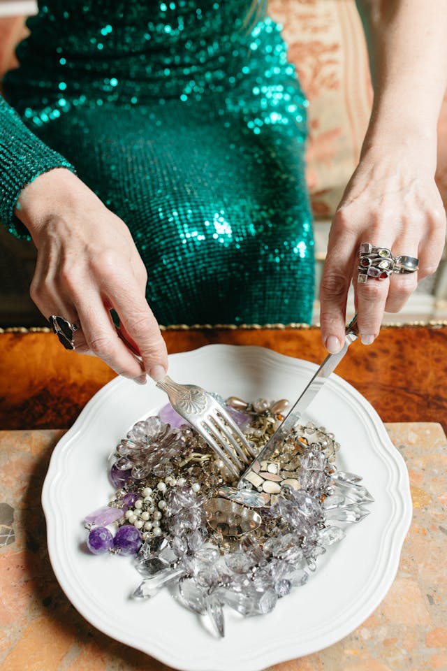 An elegantly dressed older woman in a beautiful, sequined, green dress, holding a fork and a knife, playfully attempting to eat a white ceramic plate filled with various types of jewelry.