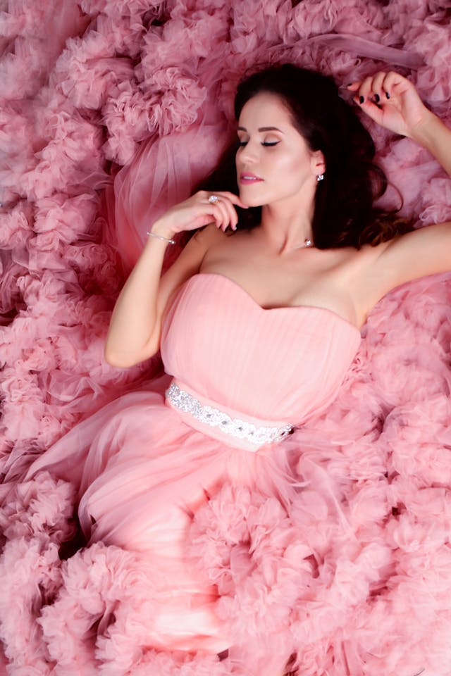 A beautiful woman in a pink dress with a sweetheart neckline, a white belt, and many layers of ruffles in it.