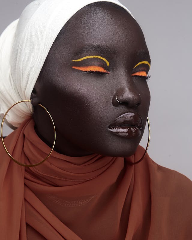 A beautiful dark skinned African woman wearing bright orange and yellow colored eyeshadow, large gold hoop earrings, a nose ring, dark brown lip gloss, a white head wrap, and a brown scarf around her neck.