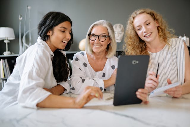 Three beautiful women sitting at a table looking at a flip board which the woman in the middle is holding in her hand.